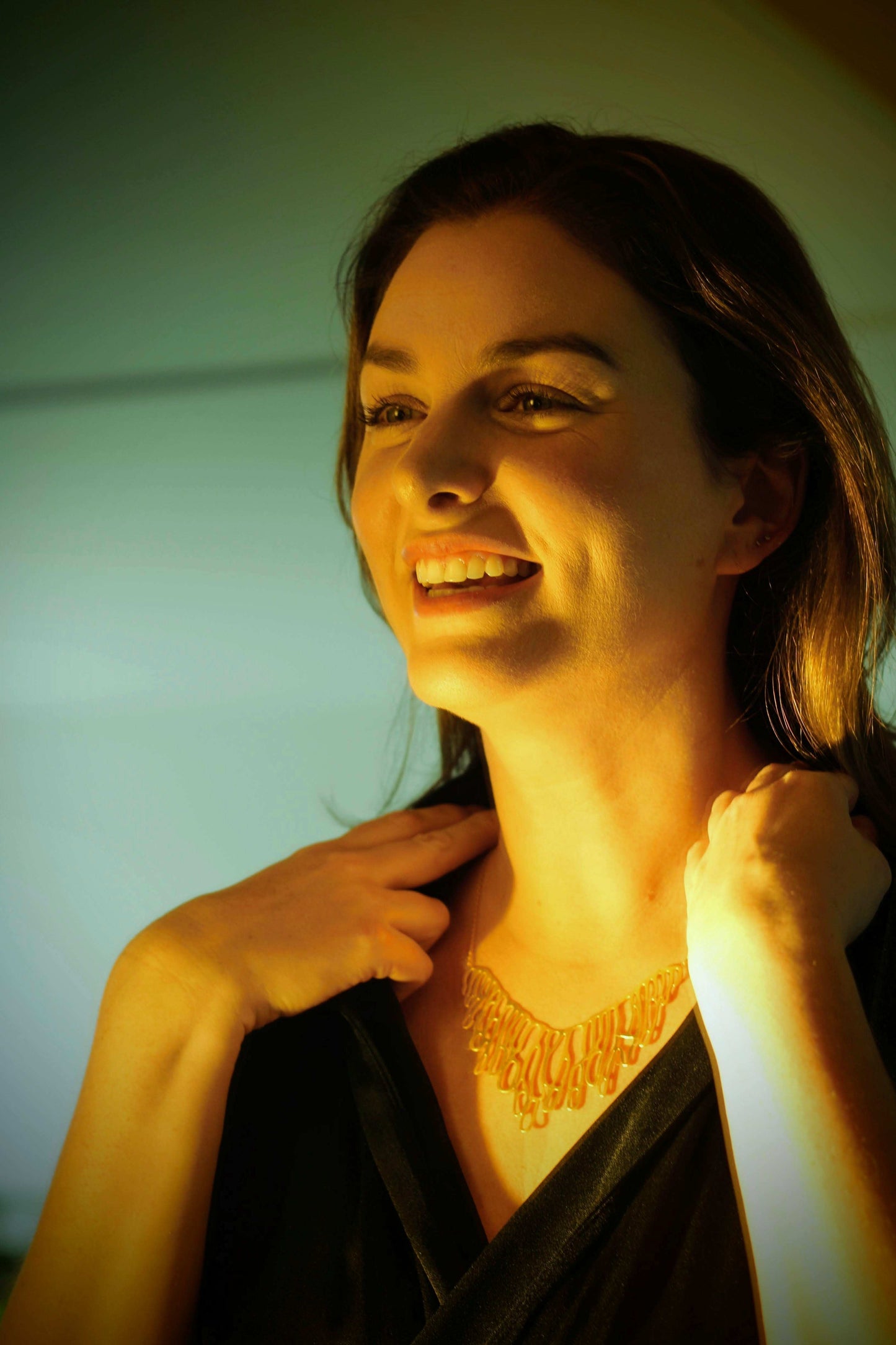 Dewdrip necklace van Olivia Peeters Master Goldsmith: Lachende vrouw met lange, donkere haren draagt een gouden halsketting met abstracte, natuurlijke, organische vormen, gefotografeerd in warm licht.