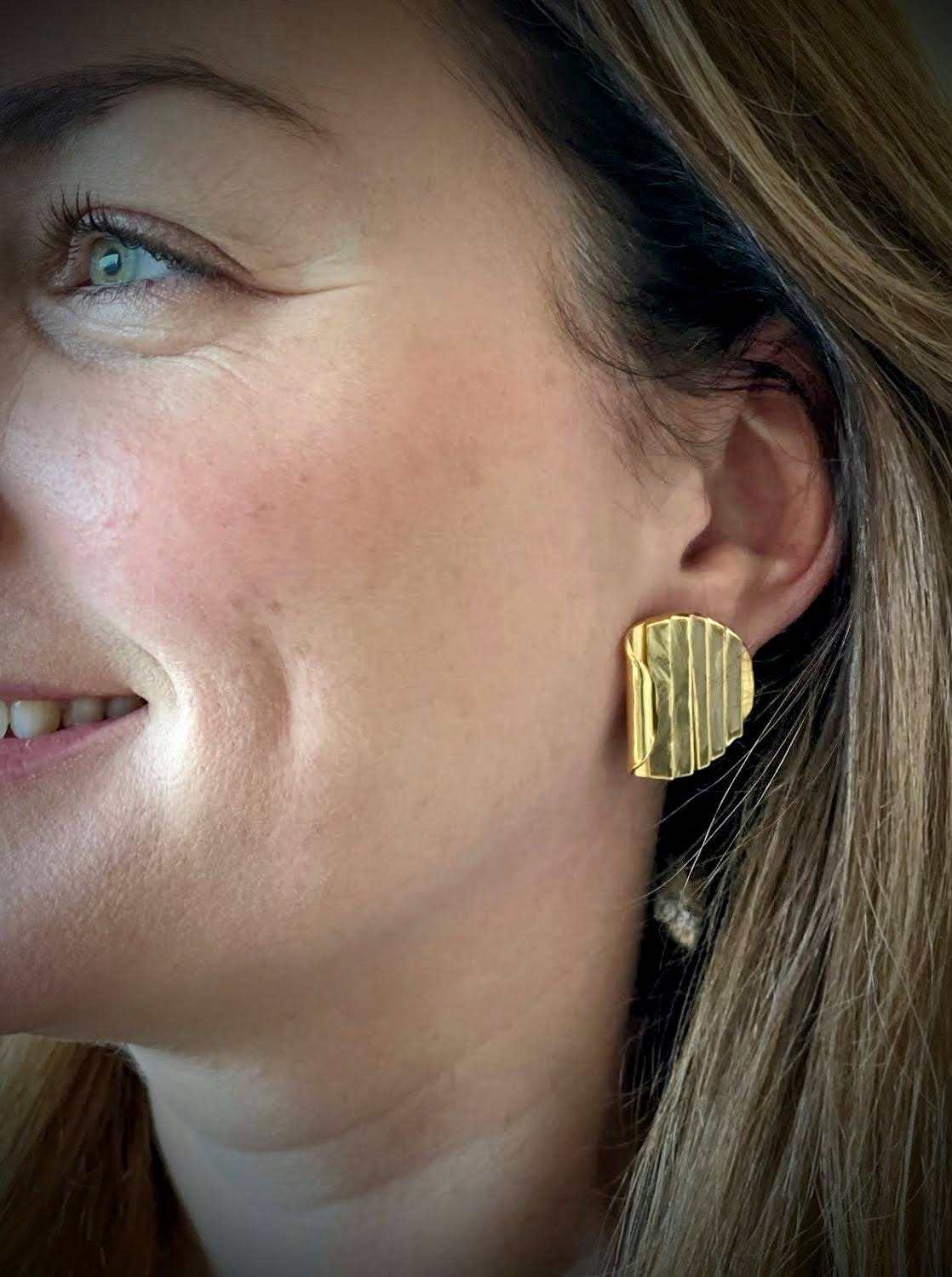 Folded Ridge Earrings van Olivia Peeters Master Goldsmith: Strakke gouden oorbellen met een geplooid, gelaagd architecturaal ontwerp, gedragen door een glimlachende vrouw met lichtbruin haar.