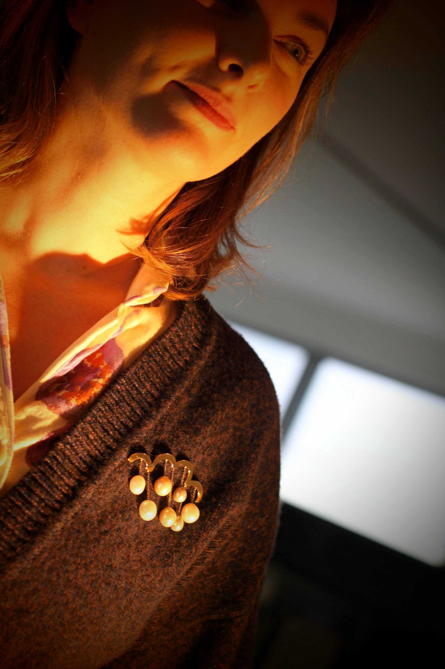 De Spring-broche van Olivia Peeters Master Goldsmith, gedragen op een donkerbruine trui door een vrouw met lang bruin haar. De broche, in de vorm van een wolk met hangende parels, wordt sfeervol verlicht door warm licht.