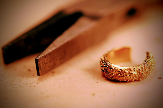Doting dots ear cuff van Olivia Peeters Master Goldsmith: Gouden ear cuff met een textuur van kleine bolletjes, liggend op een werktafel naast een tang.