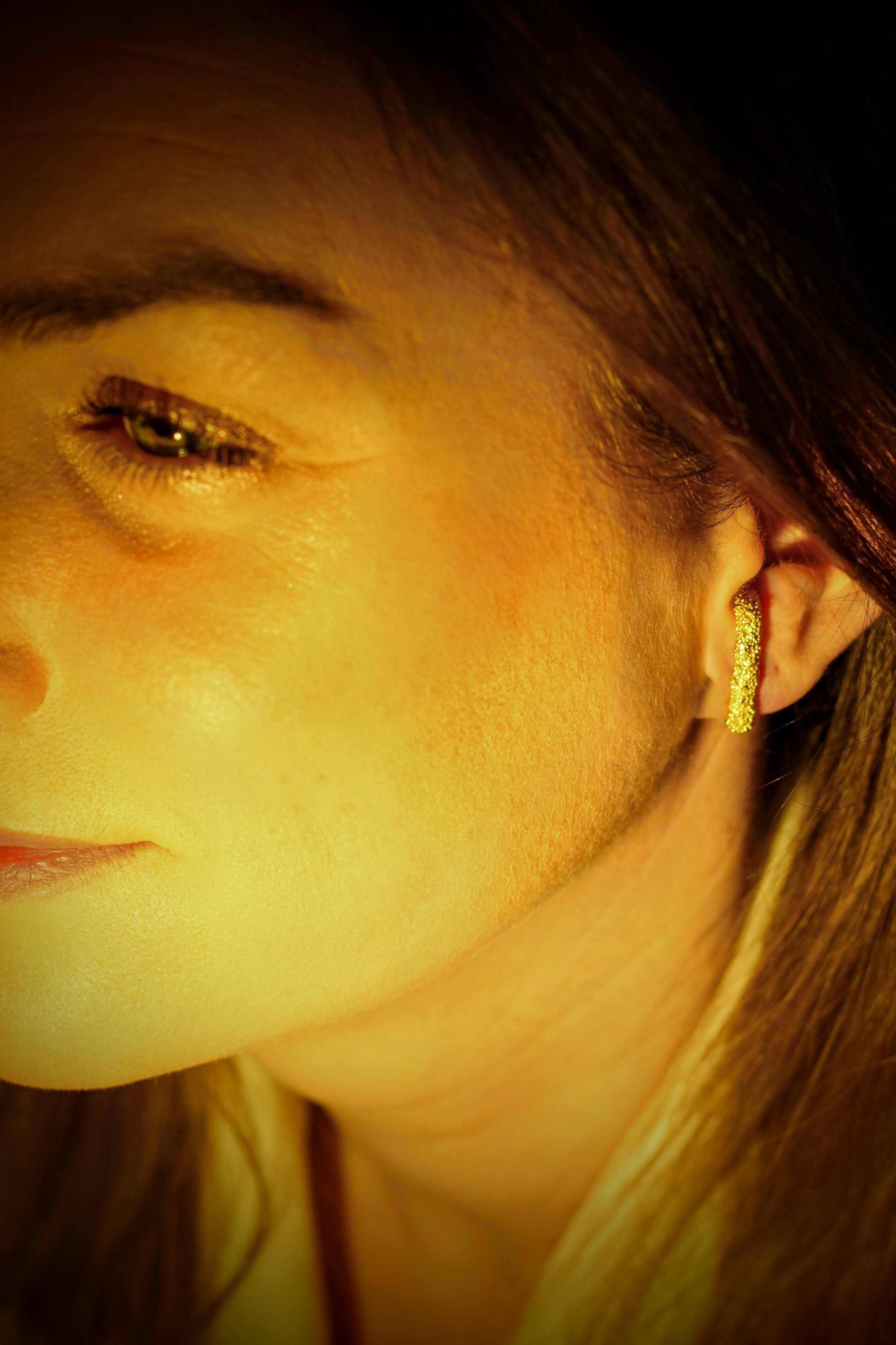 Doting dots ear suspender van olivia peeters master goldsmith: gouden stekertje met een textuur van kleine bolletjes, gedragen door een vrouw in warm licht. 