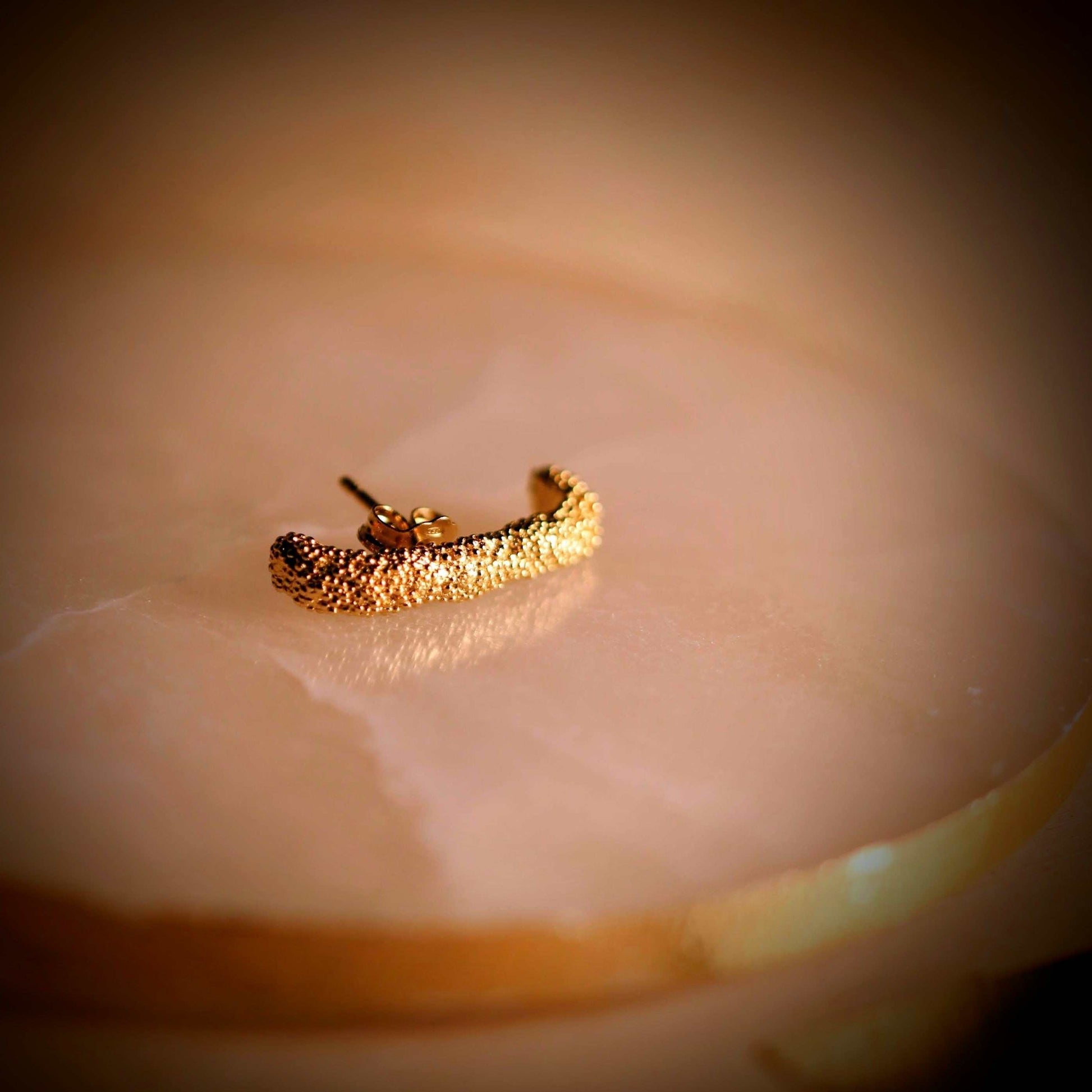 Doting dots ear suspender-oorbellen van Olivia Peeters Master Goldsmith: Gouden oorstekertje met een textuur van kleine bolletjes, liggend op een marmeren ondergrond in warm licht.