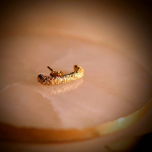 Doting dots ear suspender-oorbellen van Olivia Peeters Master Goldsmith: Gouden oorstekertje met een textuur van kleine bolletjes, liggend op een marmeren ondergrond in warm licht.