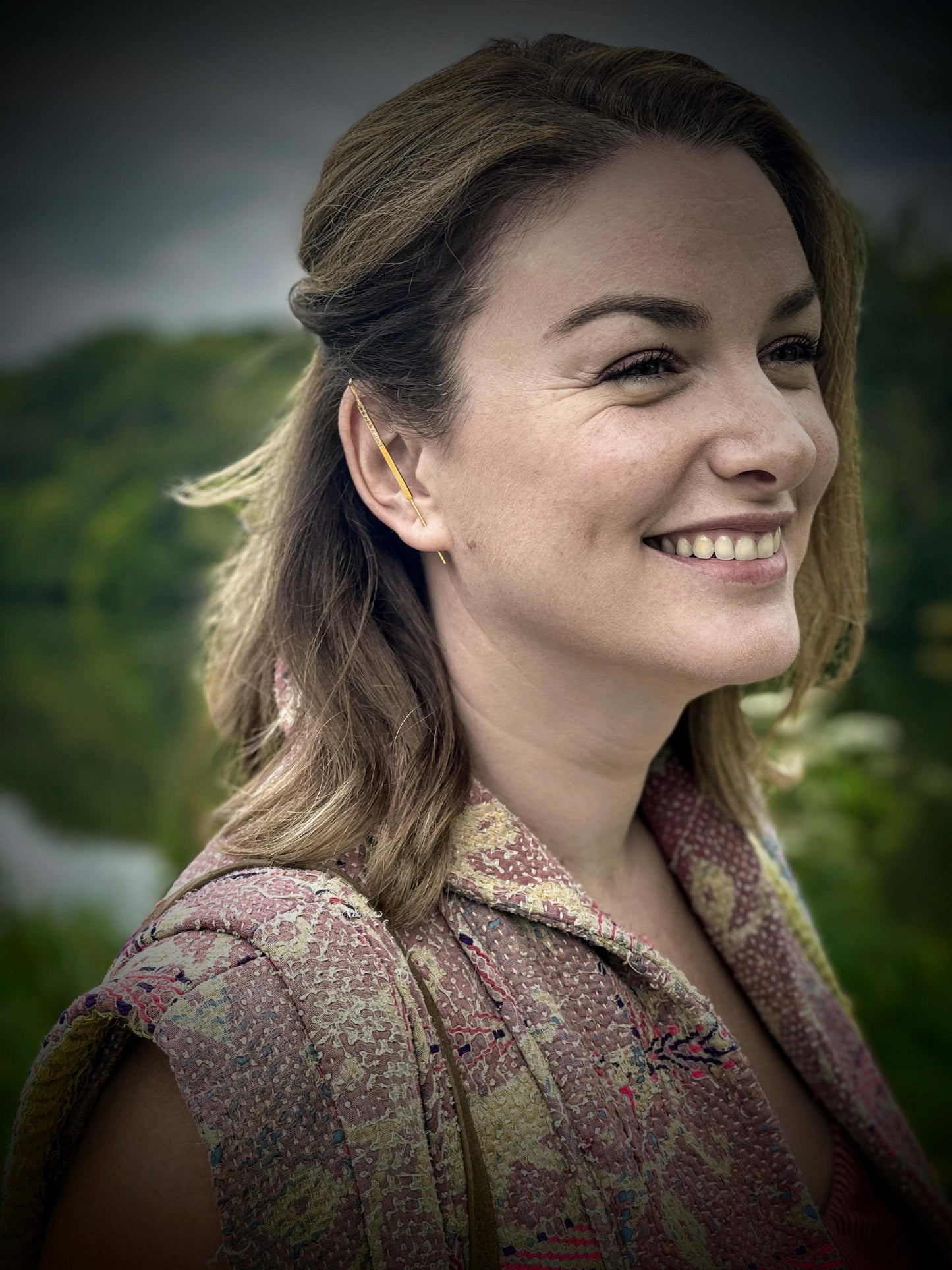 Dusk oorbel van Olivia Peeters Master Goldsmith, gedragen door een knappe, glimlachende vrouw in een kleurrijke, textuurrijke outfit, gefotografeerd in een natuurlijke buitenomgeving met groene bomen op de achtergrond. De slanke gouden oorbel loopt elegant langs de contour van het oor.