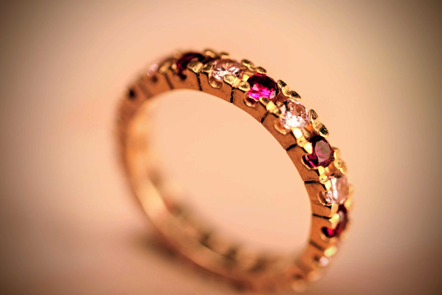 Eternal Embrace van Olivia Peeters Master Goldsmith: Elegante gouden ring met een verfijnd ontwerp, bezet met afwisselende rode en witte edelstenen, gepresenteerd tegen een zachte, neutrale achtergrond.