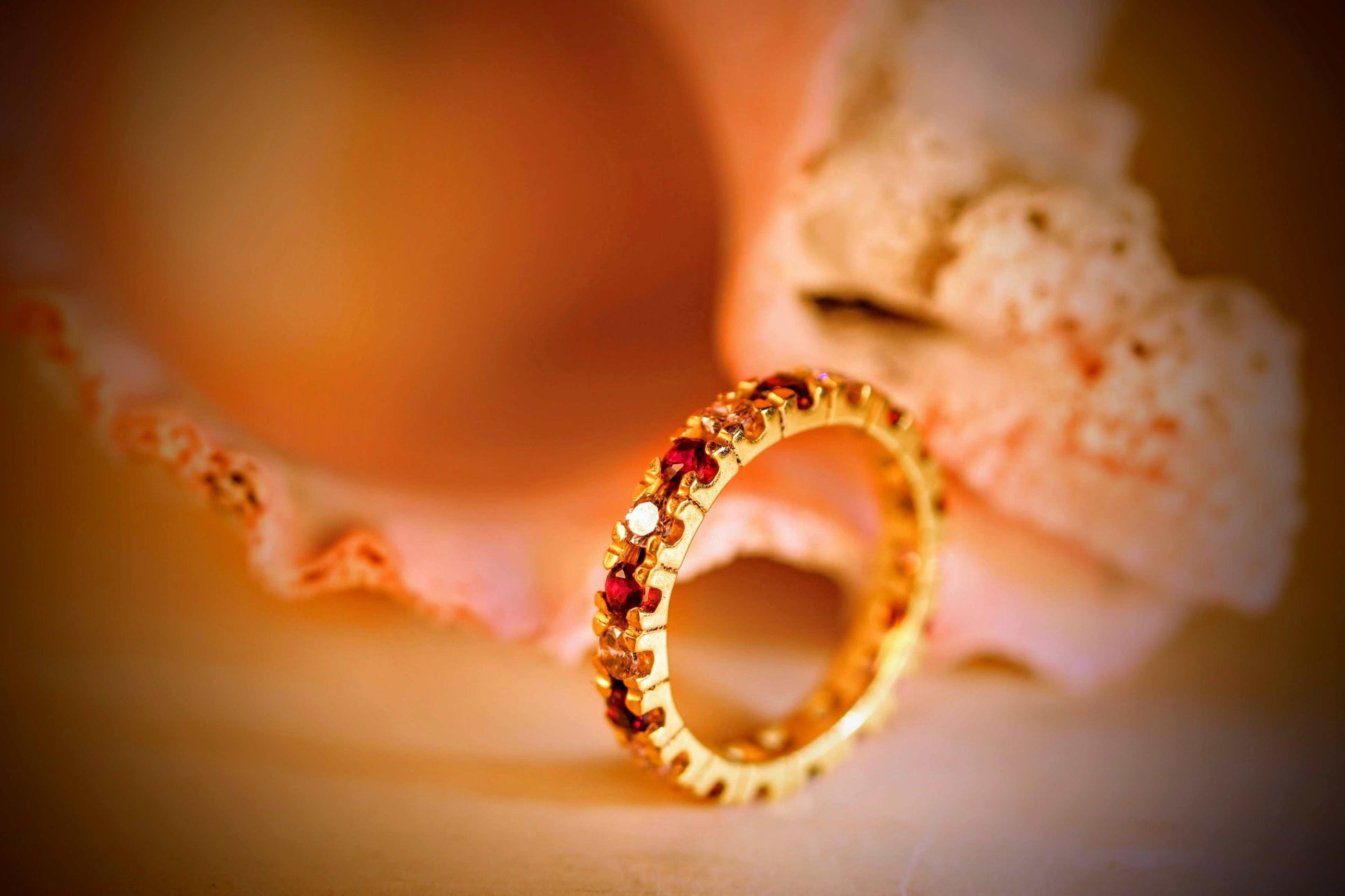Eternal Embrace van Olivia Peeters Master Goldsmith: Gouden ring met een delicate afwerking, ingelegd met rode en witte edelstenen, gepresenteerd tegen een zachte achtergrond met een zeeschelp in warm licht.