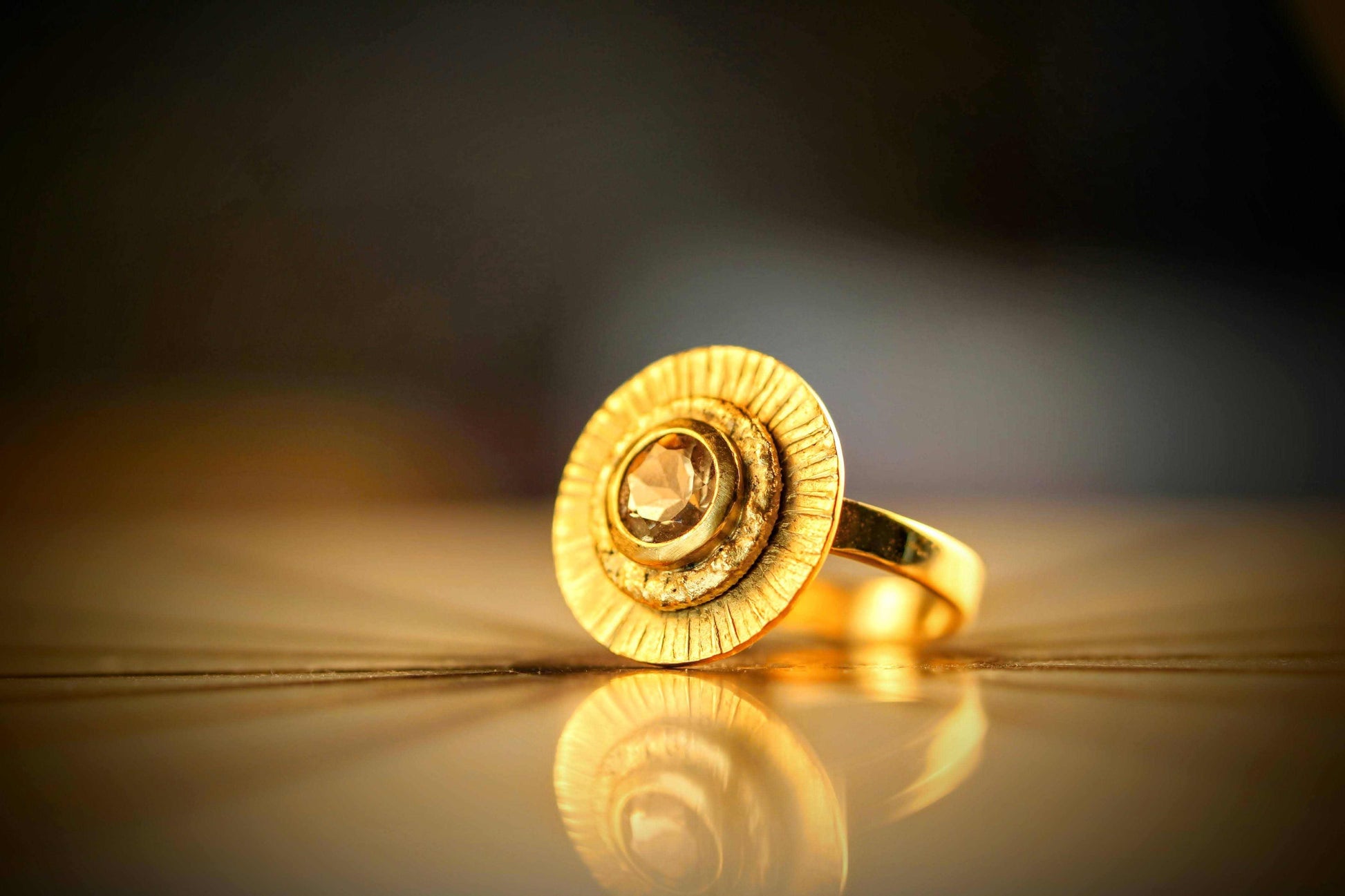 Fall Ring van Olivia Peeters Master Goldsmith: Gouden ring met een unieke ronde ontwerp, voorzien van een centrale bruine diamant met een facettenpatroon, omlijst door subtiele textuurdetails. De ring ligt op een reflecterende ondergrond, gevangen in warm en sfeervol licht.