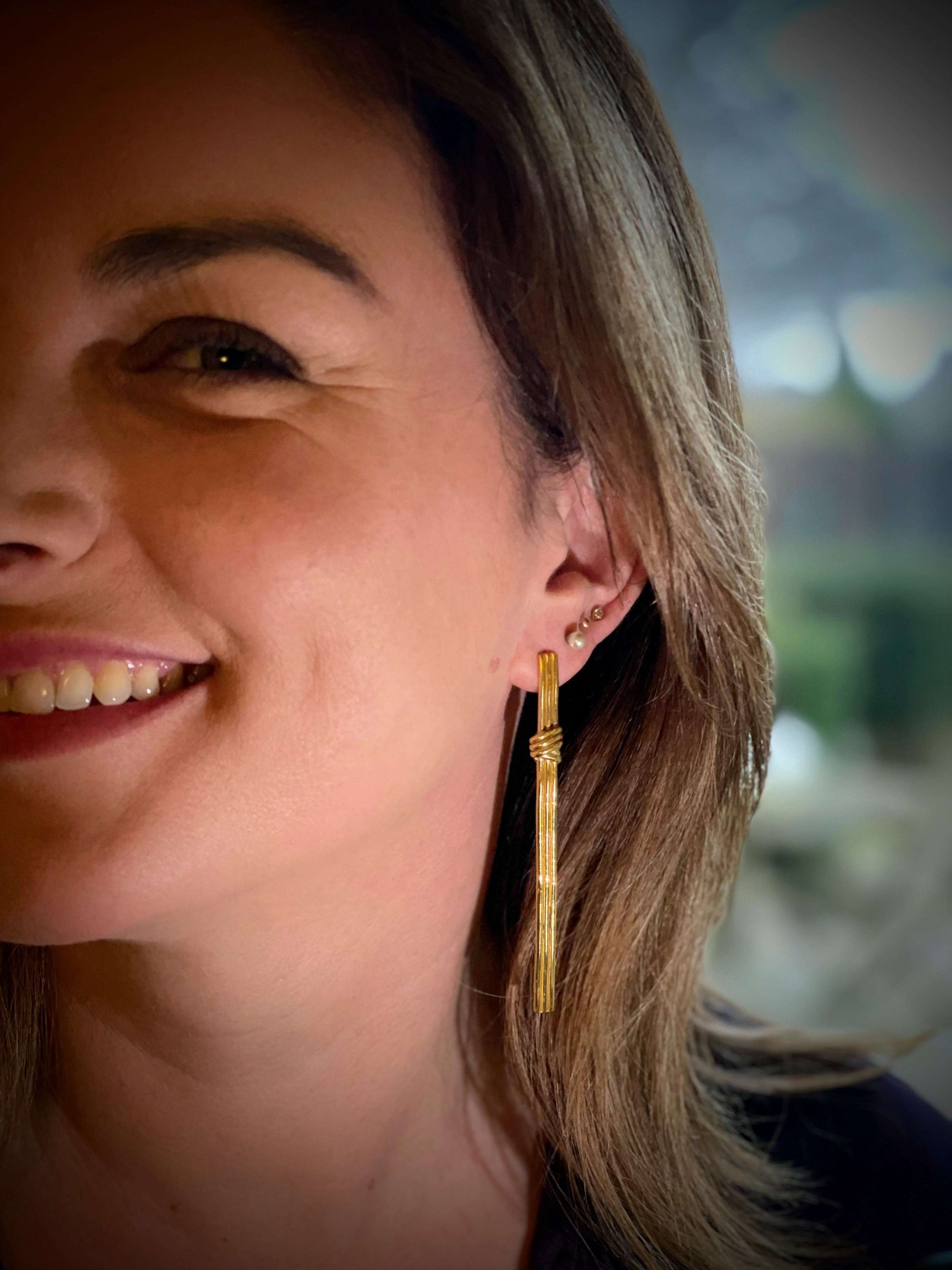 Gordian Knot Earrings van Olivia Peeters Master Goldsmith: een gouden lange strakke oorbel met wikkeldetails, gedragen door een lachende vrouw met bruine haren..