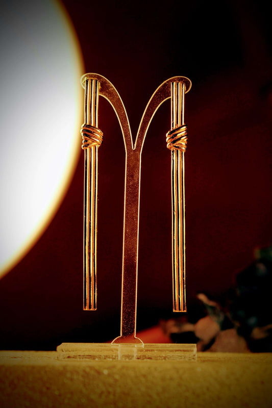 Gordian Knot Earrings van Olivia Peeters Master Goldsmith: Gouden lange strakke oorbellen met een minimalistisch ontwerp en wikkeldetails, gepresenteerd op een sieradendisplay in warm licht.