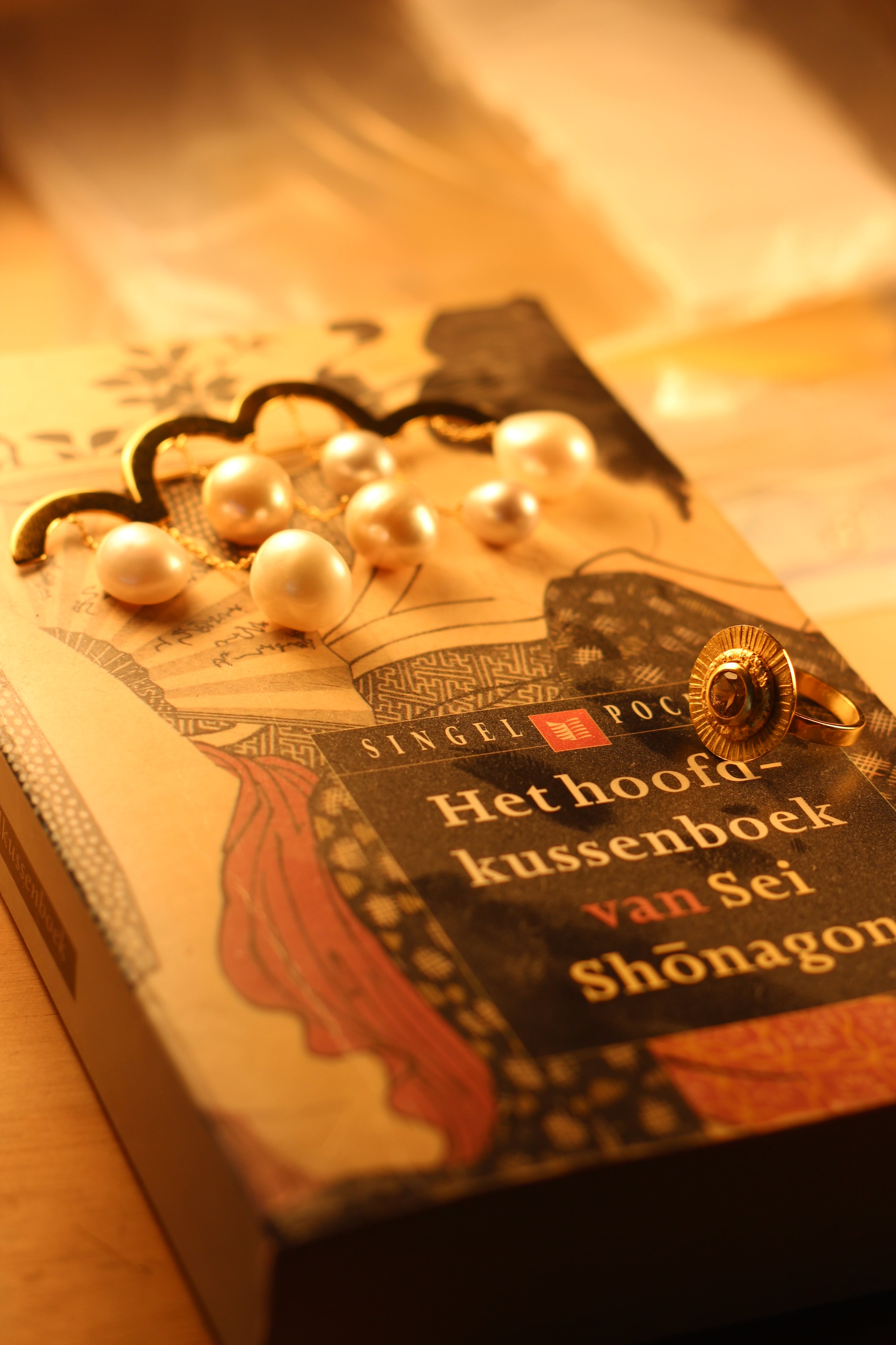 Een sfeervolle compositie met het boek "Het hoofdkussenboek van Sei Shōnagon" in een warme, gouden gloed, vergezeld van een elegante gouden ring en een broche met parels, die een klassieke en tijdloze uitstraling oproepen.