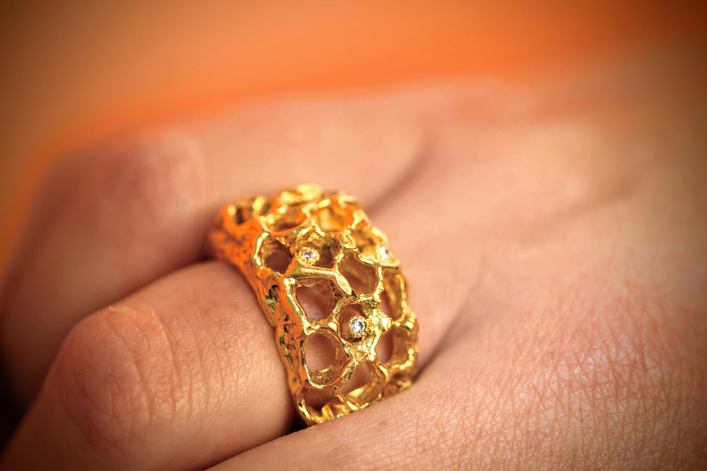 Honeycomb van Olivia Peeters Master Goldsmith: Gouden ring met een opengewerkt honingraatdesign en subtiele diamantjes, gedragen aan een vinger tegen een zachte, warme achtergrond.