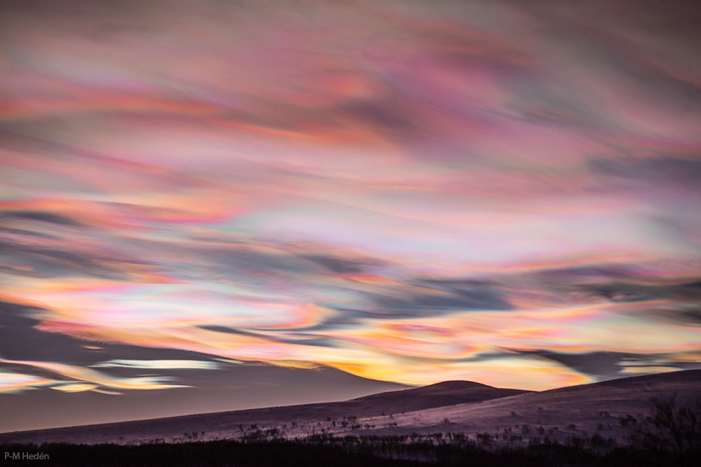 Iriserende wolken tijdens een zonsondergang, met zachte pastelkleuren in roze, paars en geel die een surrealistisch en etherisch landschap creëren boven een glooiend terrein.