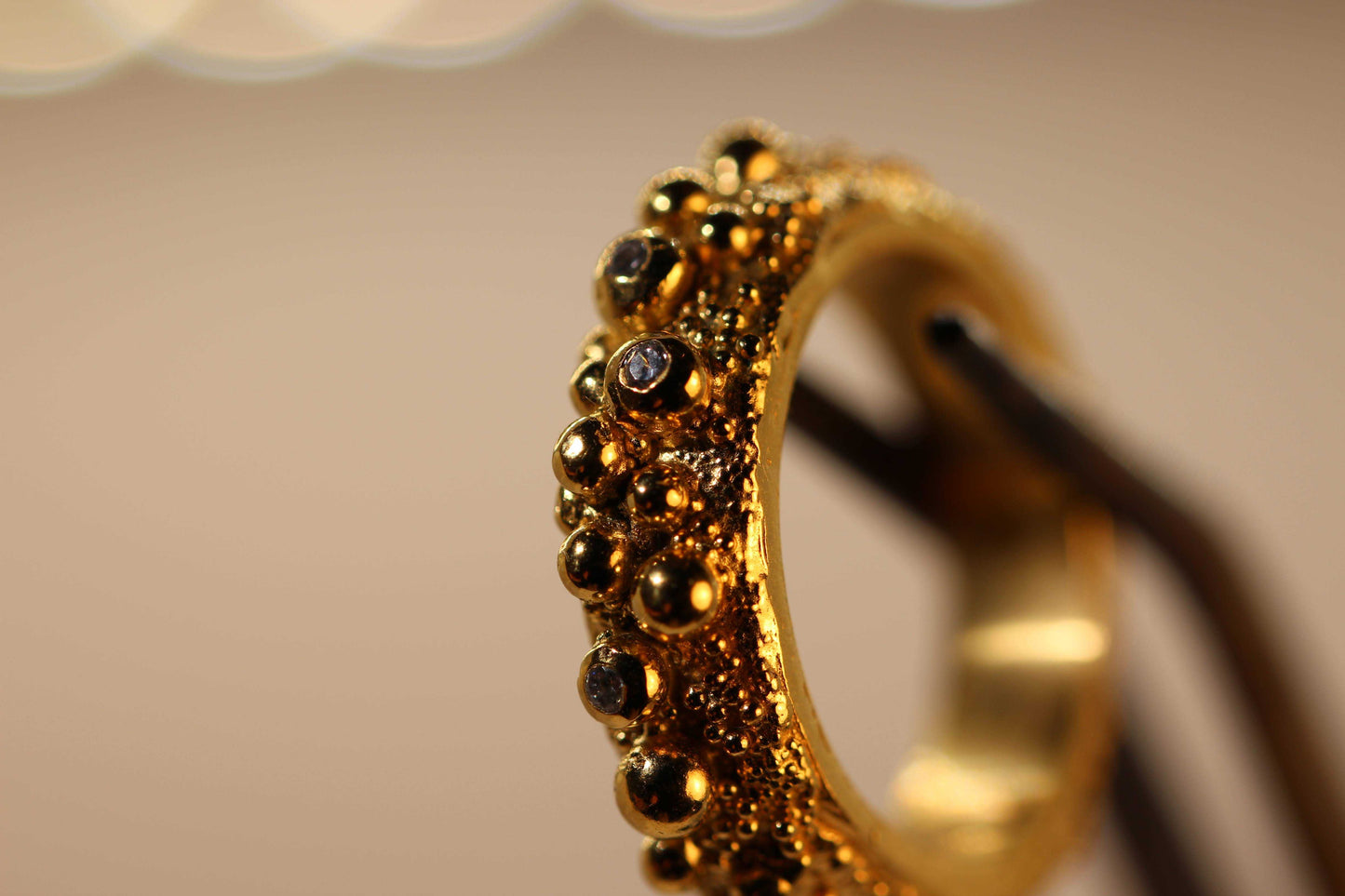 Love Bubbles ring van Olivia Peeters Master Goldsmith, een unieke gouden ring met speelse bolvormige details en subtiele donkere accenten, gestileerd op een houder tegen een neutrale achtergrond om de complexe textuur en glans te benadrukken.