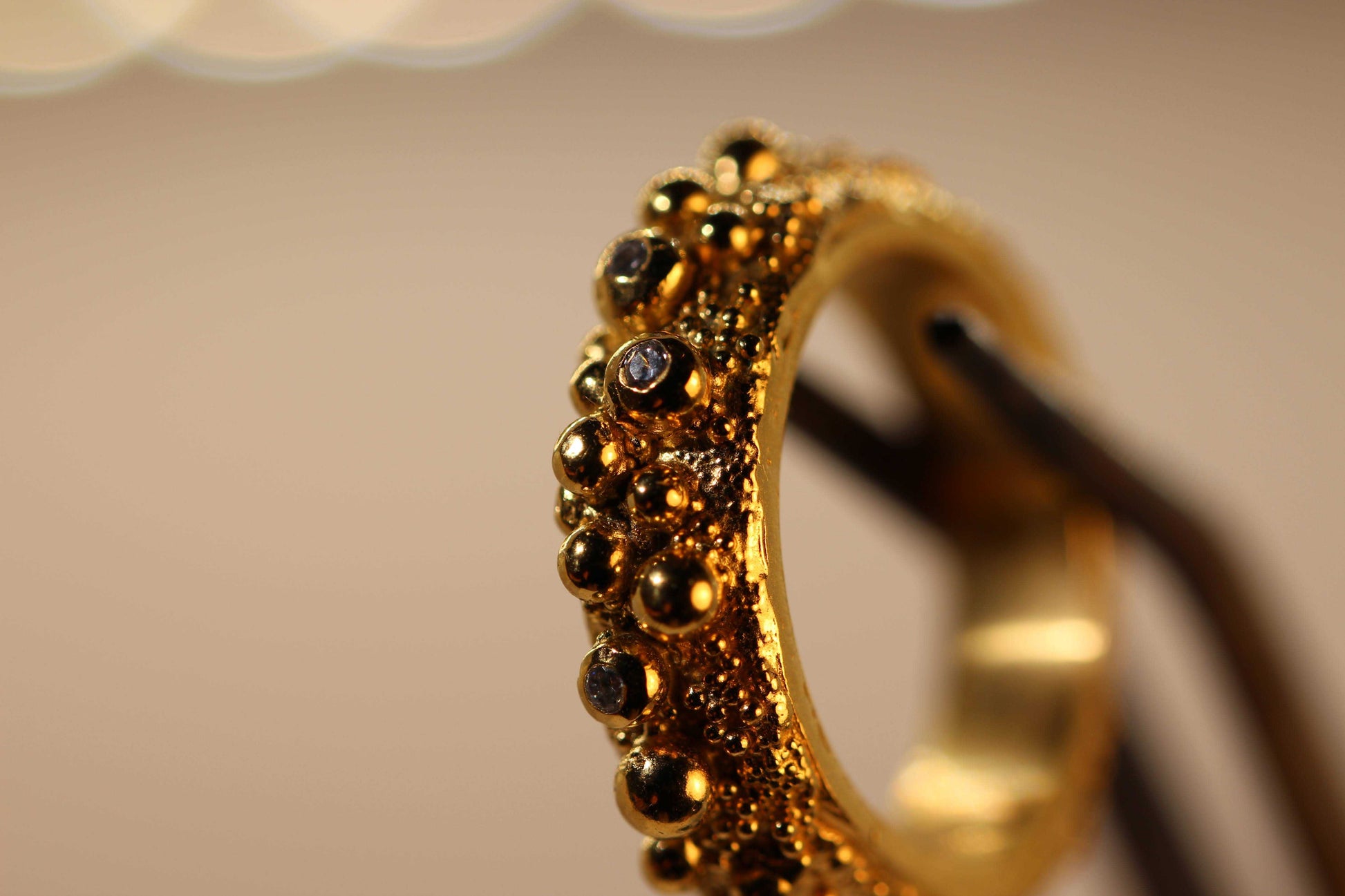 Love Bubbles ring van Olivia Peeters Master Goldsmith, een unieke gouden ring met speelse bolvormige details en subtiele donkere accenten, gestileerd op een houder tegen een neutrale achtergrond om de complexe textuur en glans te benadrukken.