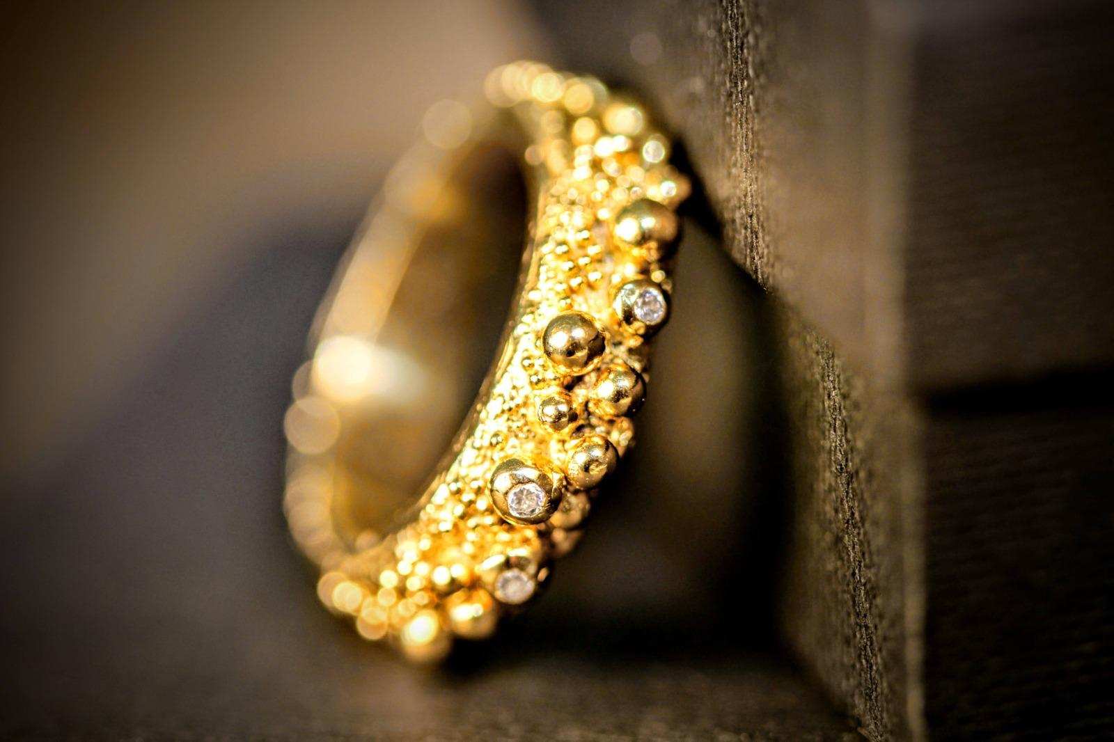 Love Bubbles ring van Olivia Peeters Master Goldsmith, close-up van een gouden ring met een speels en organisch ontwerp van verschillende maten gouden bolletjes, waarin kleine fonkelende diamanten zijn verwerkt, afgebeeld tegen een donkere, sfeervolle achtergrond.