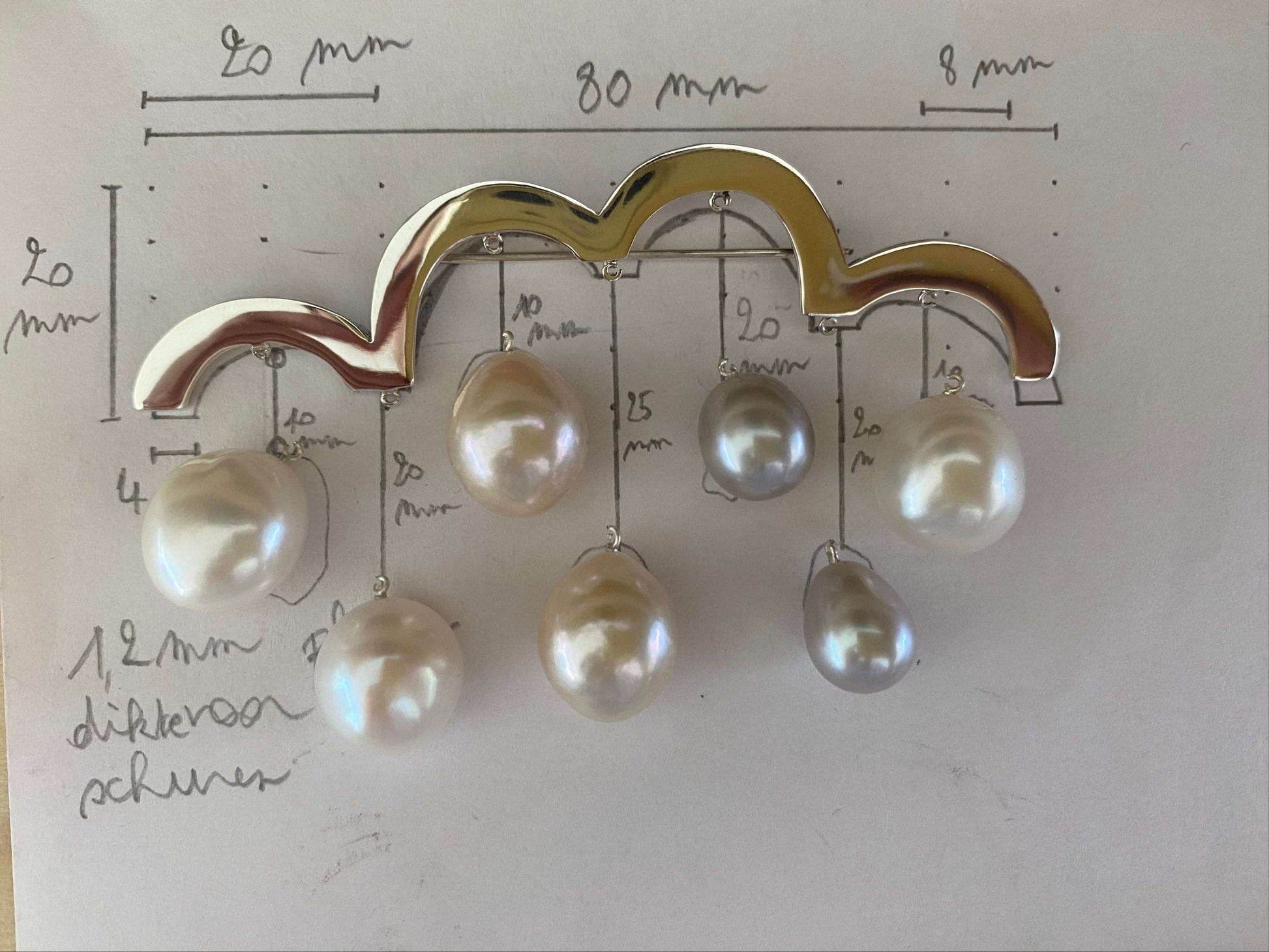 Spring Brooch van Olivia Peeters Master Goldsmith: een gouden broche in de vorm van een wolk met daaraan hangende parels in verschillende maten en tinten, geplaatst op een schets met technische afmetingen en notities over het ontwerp.