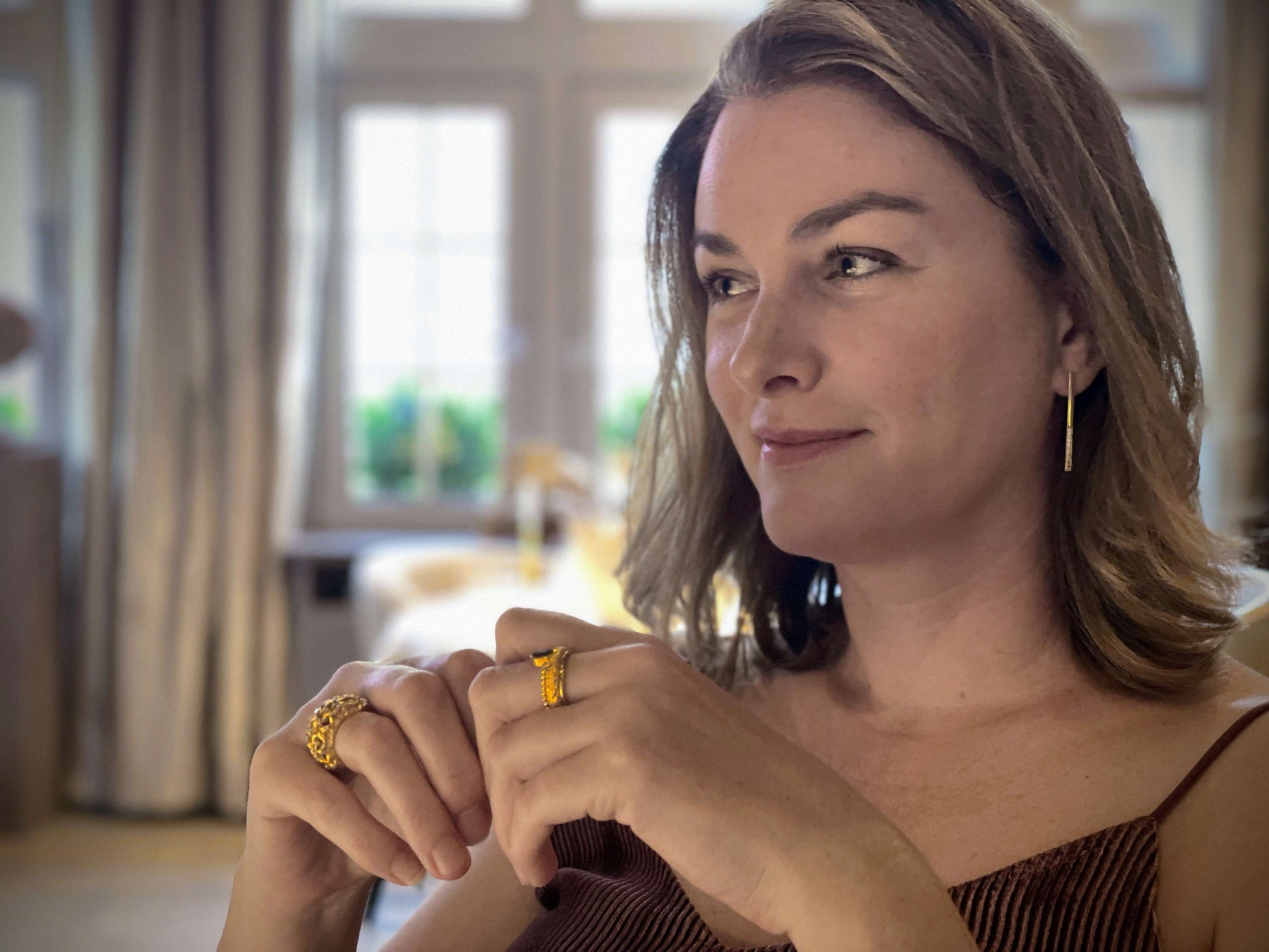 Een knappe vrouw draagt sieraden van Olivia Peeters Master Goldsmith, waaronder de Dusk & Dawn oorbellen, de Honeycomb Ring en de Stellwagen Ring, terwijl ze ontspannen poseert in een elegante kamer met een zachte achtergrond.