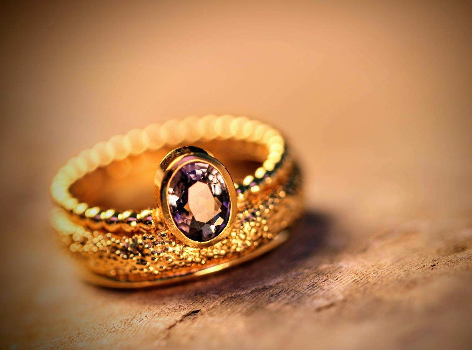 Stellwagen Ring van Olivia Peeters Master Goldsmith, een elegante gouden ring met een verfijnd kralenpatroon en een opvallende ovale paarse edelsteen in een gepolijste zetting, weergegeven tegen een warme houten achtergrond.