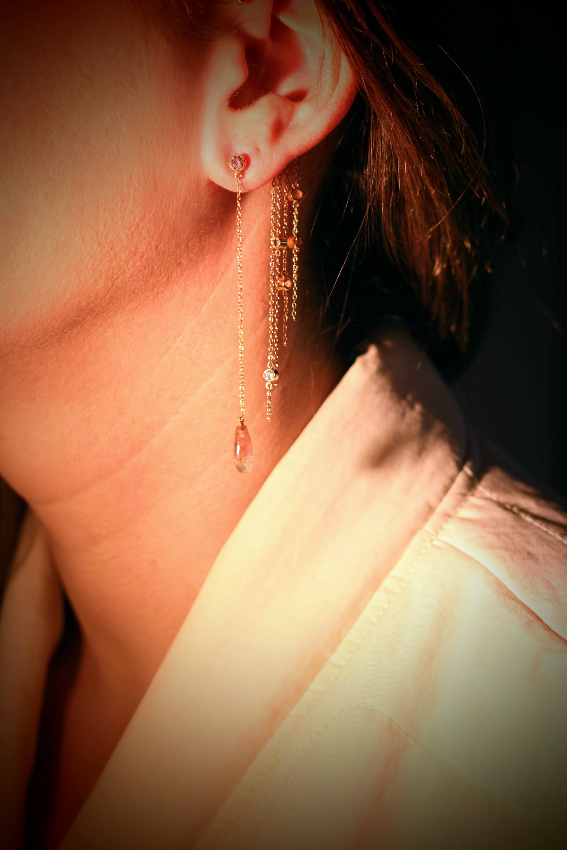 Summer earrings van Olivia Peeters Master Goldsmith: een elegant oor dat een lange gouden oorbel draagt met fijne kettingen, bezet met kristallen en een druppelvormige hanger. Het licht accentueert de glans van het goud en de delicate details van de oorbel.