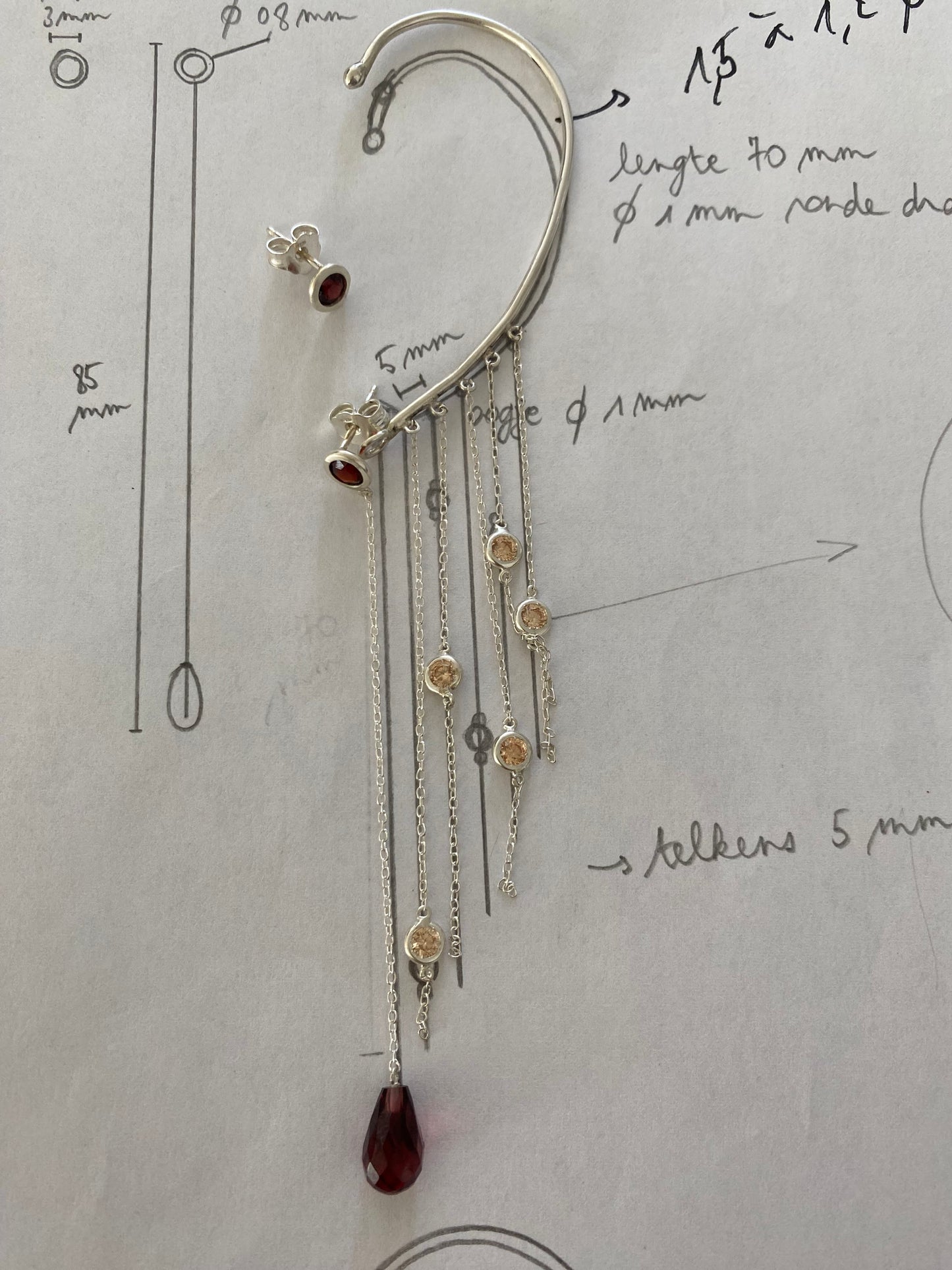 Een schetsblad met technische tekeningen waarop een handgemaakte zilveren oorbel is geplaatst. De oorbel heeft een gebogen vorm met meerdere fijne kettingen die eindigen in cirkelvormige zettingen en een druppelvormige rode hanger. De schets toont afmetingen en ontwerprichtlijnen.