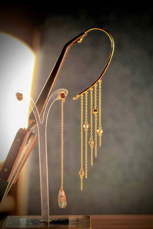 Summer-oorbellen van Olivia Peeters Master Goldsmith. Een delicate combinatie van gouden kettingen met ingelegde edelstenen en een druppelvormige hanger, gepresenteerd op een stijlvolle houder tegen een subtiele achtergrond.