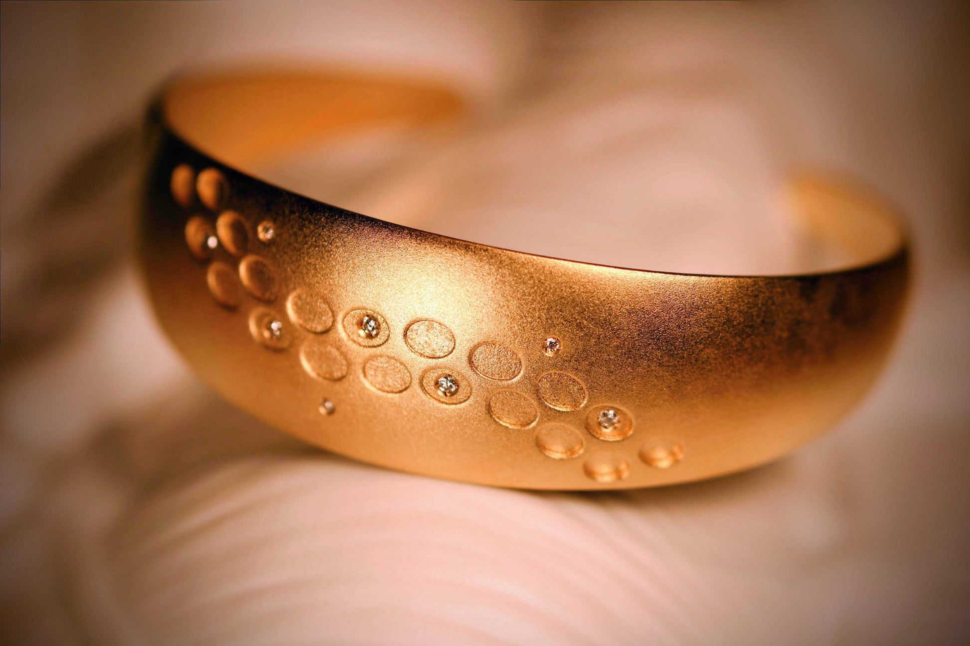 Close-up van een brede gouden armband met een subtiele matte afwerking. De armband is versierd met een patroon van gegraveerde cirkels en ingelegde diamanten, wat een verfijnde en luxe uitstraling geeft.