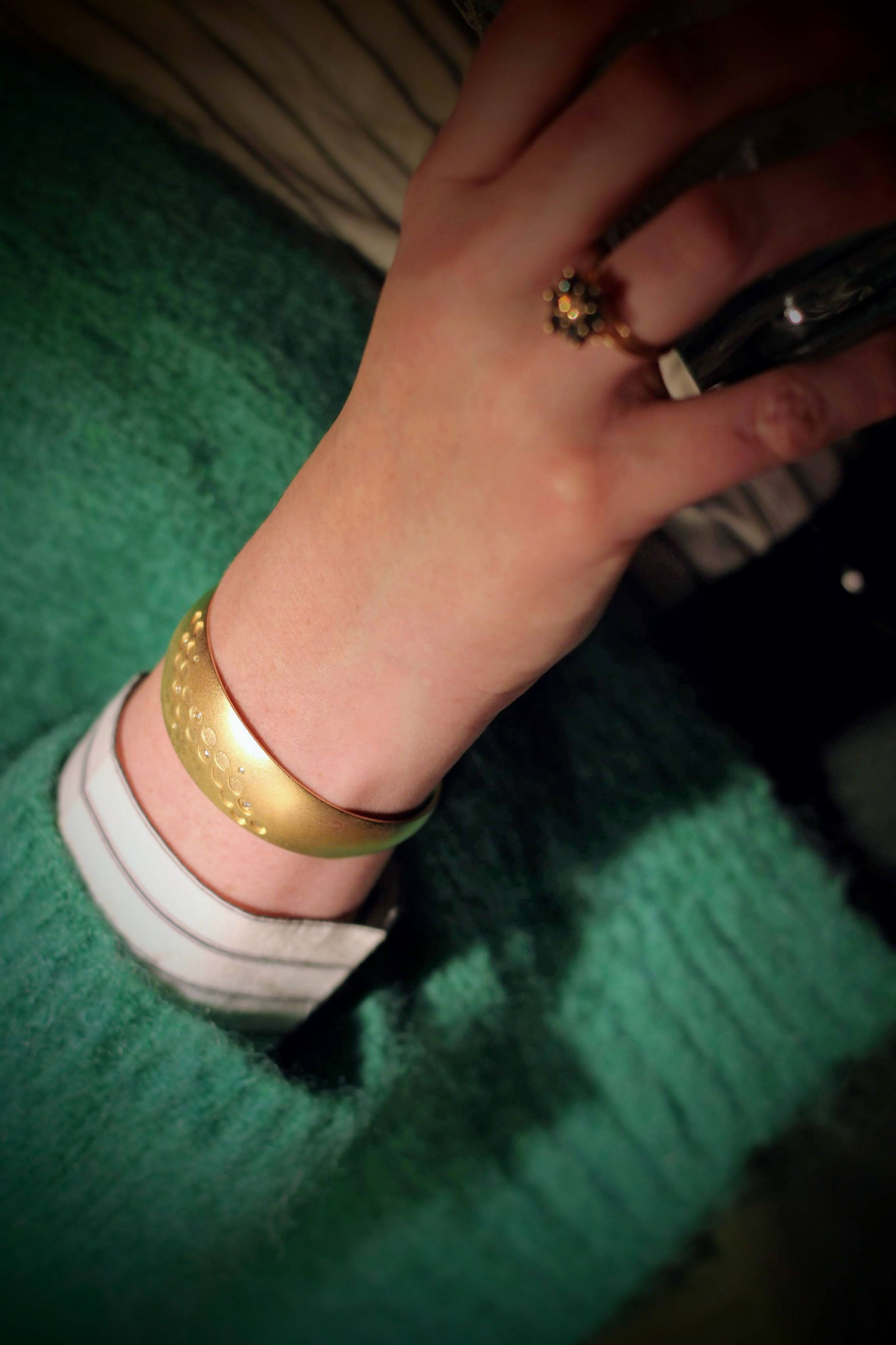 Een gouden armband met een matte afwerking en cirkelpatroon wordt gedragen om de pols van een persoon, gecombineerd met een groene trui en een elegante gouden ring met een bloemvormig design.