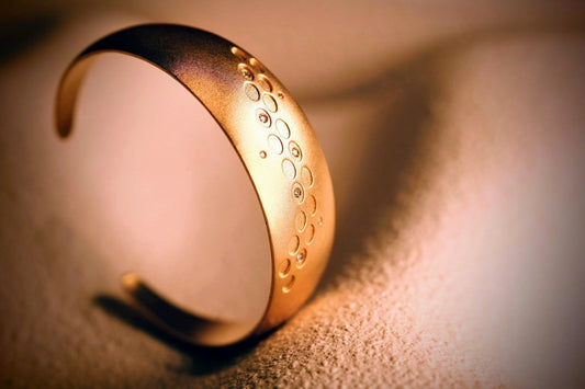 Winter Bracelet van Olivia Peeters Master Goldsmith, een matte, gebogen gouden armband met een patroon van cirkels en ingelegde diamanten die sprankelen in het zachte licht.