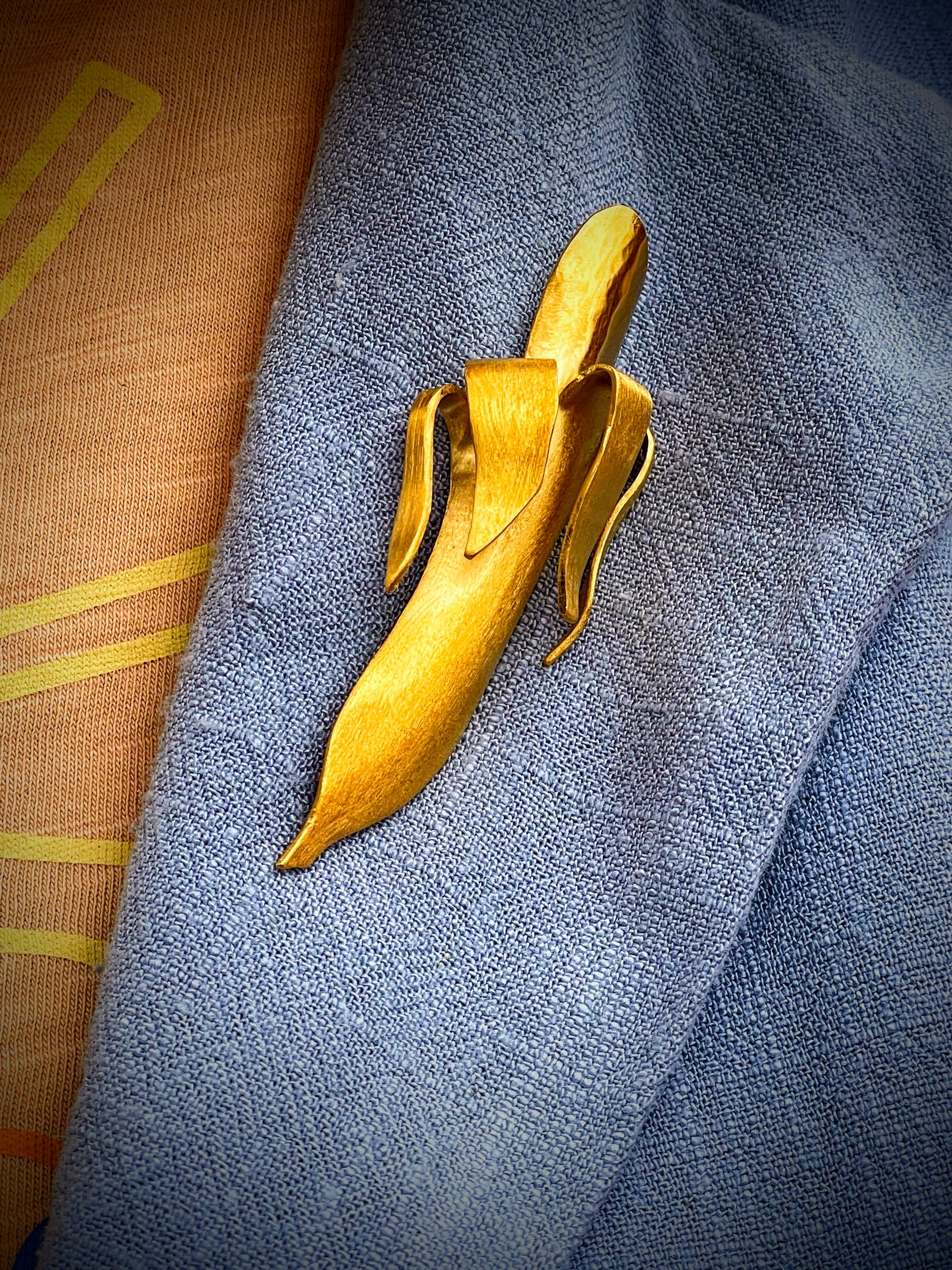 Banana Brooch van Olivia Peeters Master Goldsmith: Gouden broche in de vorm van een abstracte banaan met een matte afwerking, gepresenteerd op een blauwe stoffen vest.