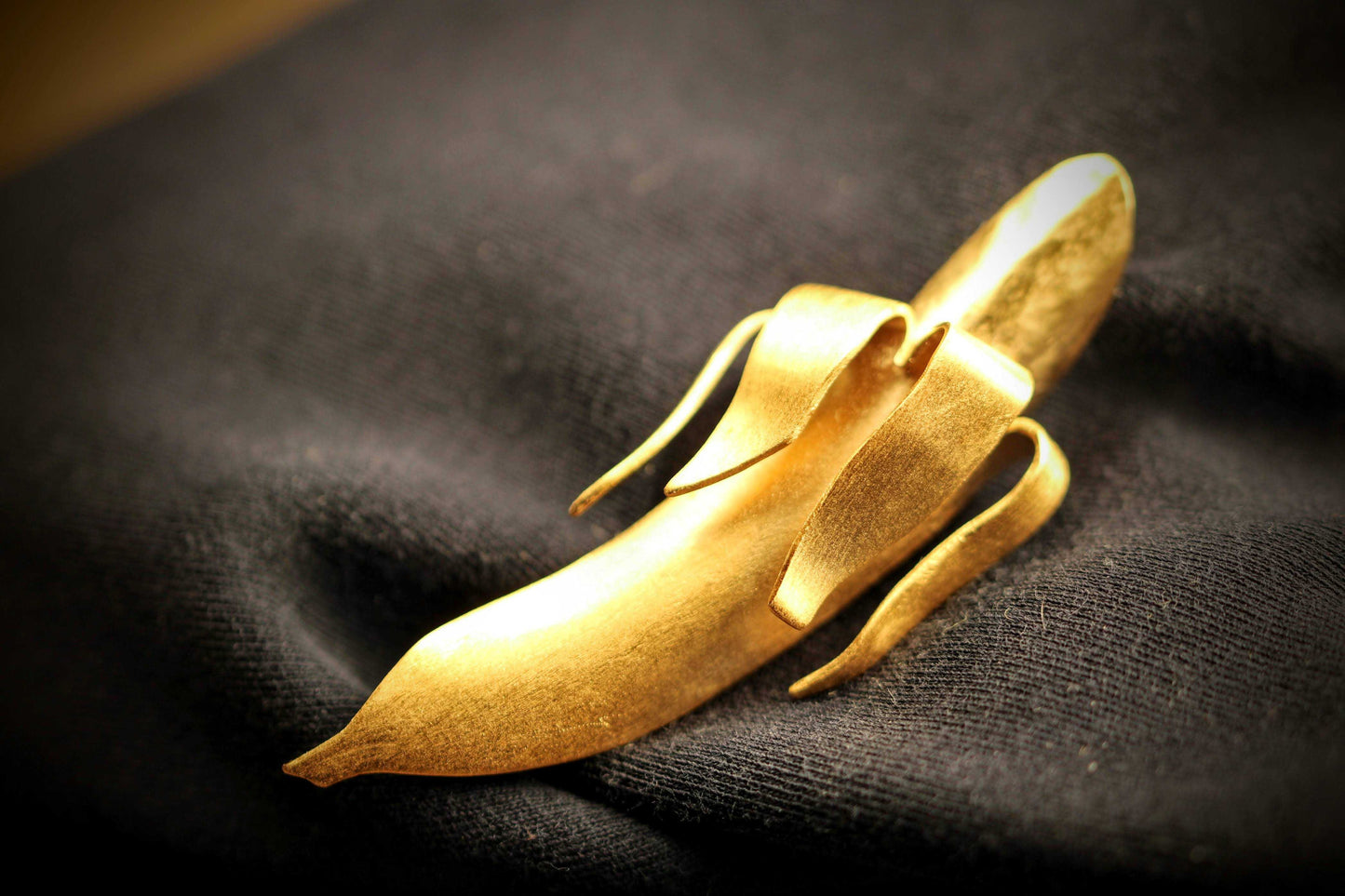 Banana Brooch: broche van Olivia Peeters Master Goldsmith: Gouden broche in de vorm van een abstracte banaan met elegante lijnen en een matte afwerking, gepresenteerd op een donkere textielachtergrond in zacht licht.