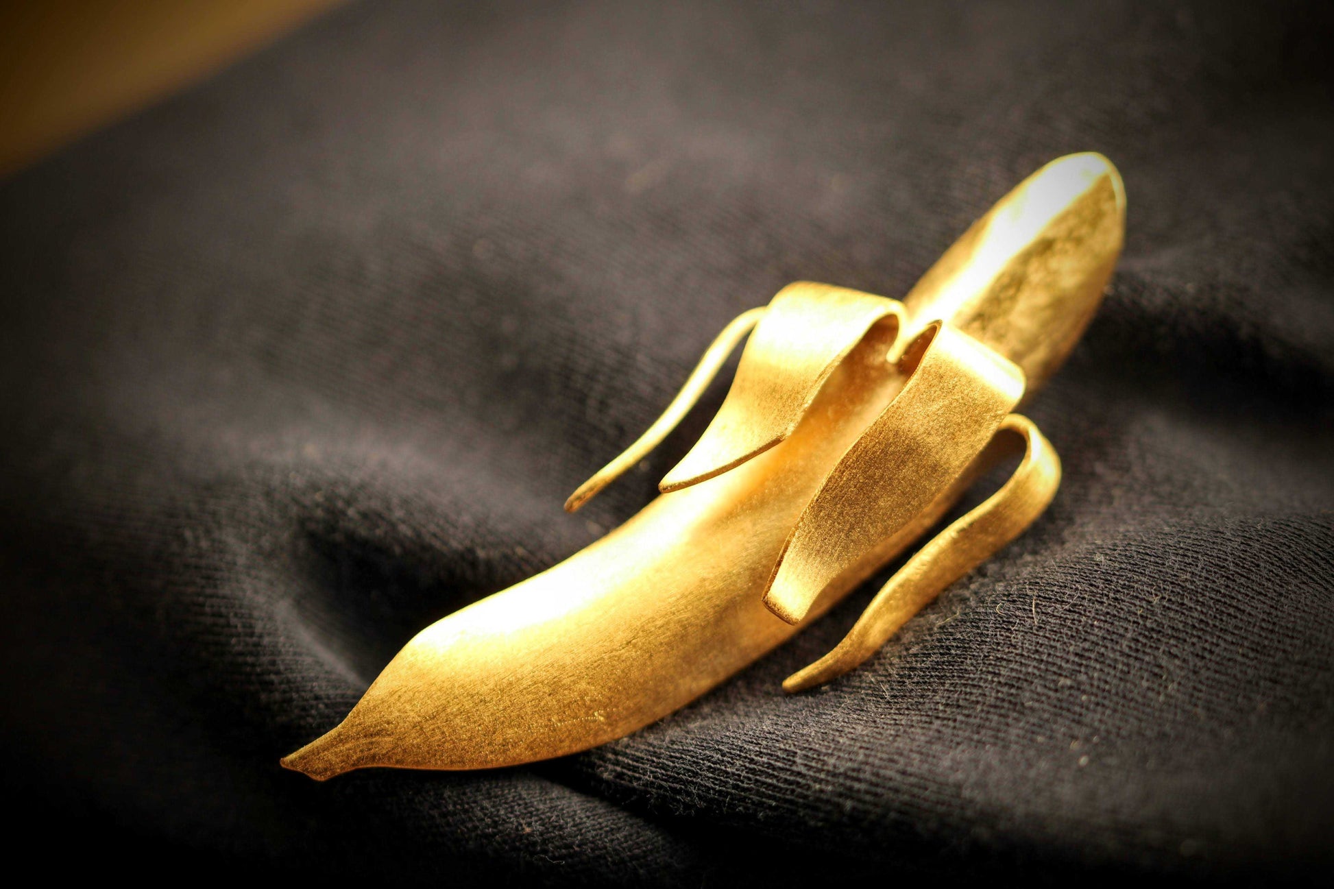 Banana Brooch: broche van Olivia Peeters Master Goldsmith: Gouden broche in de vorm van een abstracte banaan met elegante lijnen en een matte afwerking, gepresenteerd op een donkere textielachtergrond in zacht licht.
