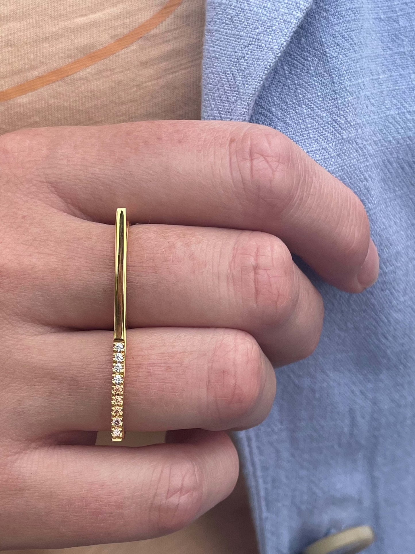 Morning Horizon van Olivia Peeters Master Goldsmith: Gouden ring met een modern en minimalistisch ontwerp, voorzien van een rechte balk en een lijn van fonkelende diamanten, gedragen aan een hand tegen een lichtblauwe en beige achtergrond.