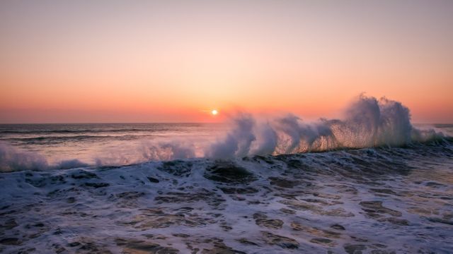 Een prachtige zonsondergang boven een onrustige oceaan met golven die breken en schuim vormen, wat de dynamiek van de natuur vastlegt tegen een serene hemel in zachte roze en oranje tinten.