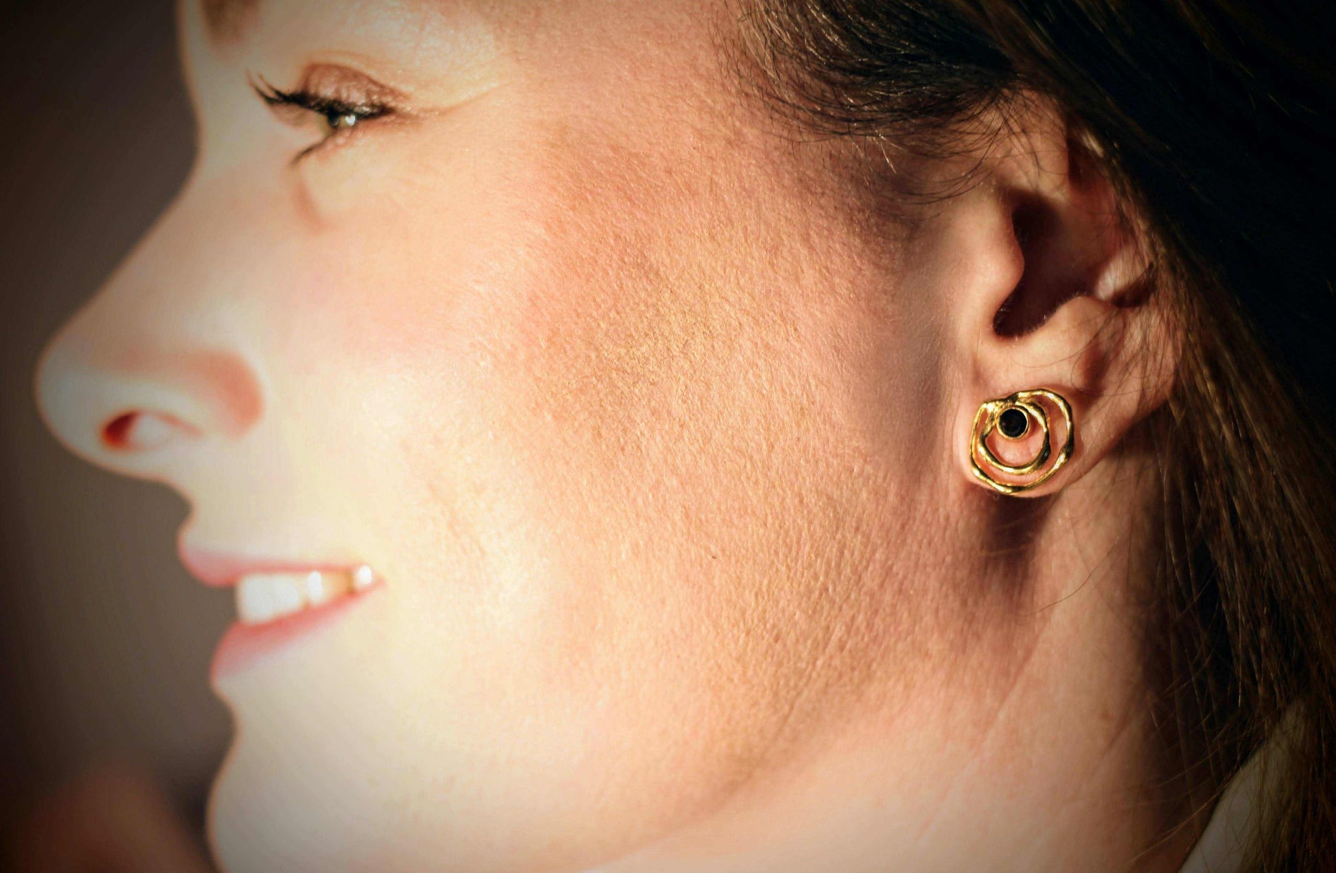 Ripple Drop-studoorbellen van Olivia Peeters Master Goldsmith, gedragen door een mooie vrouw. Gouden oorringen met een organisch golvend ontwerp en een donkere edelsteen als accent.