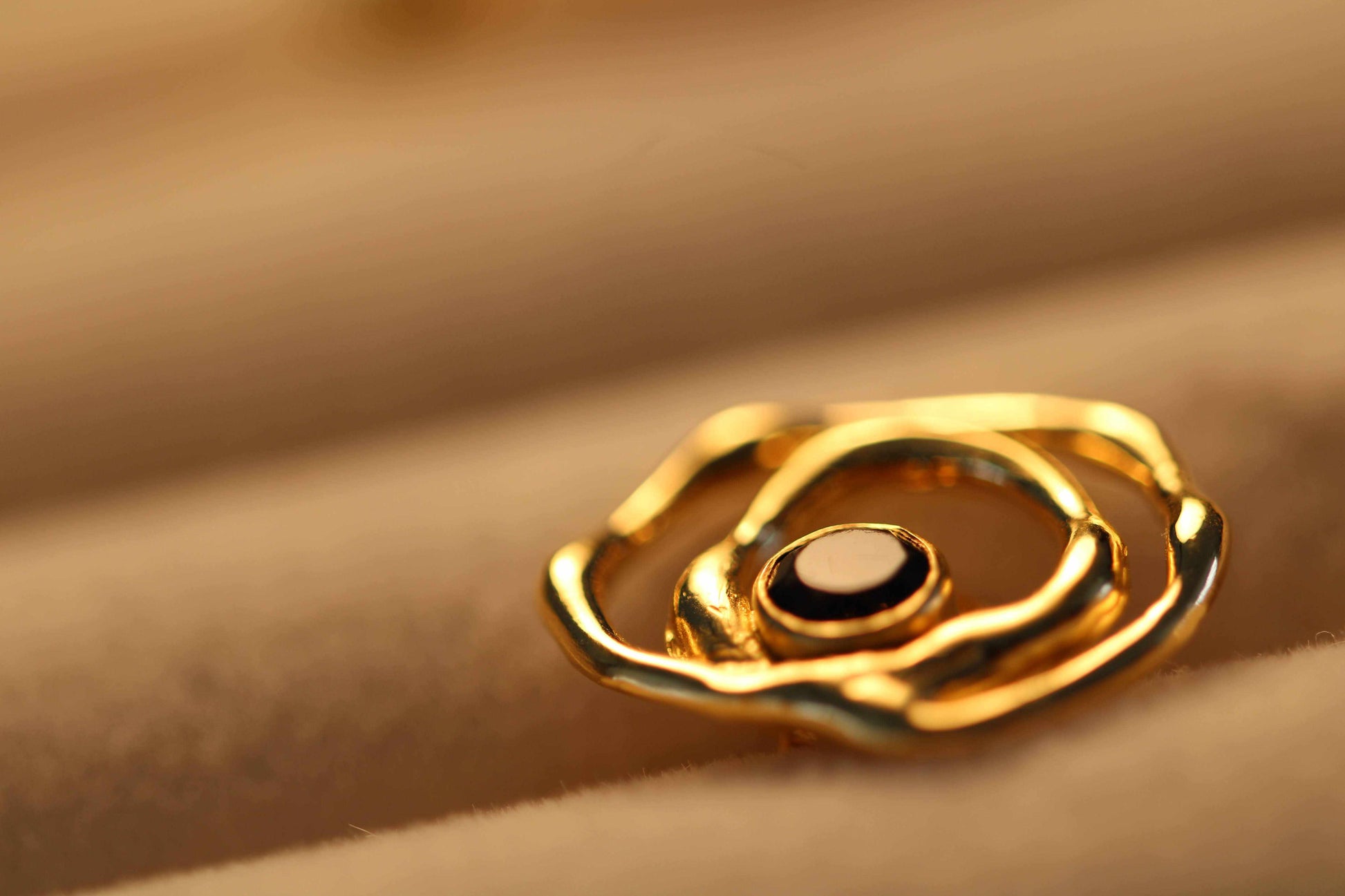 Ripple Drop-studoorbellen van Olivia Peeters Master Goldsmith. Close-up van een gouden oorbel met een organisch, golvend ontwerp en een donkere edelsteen in het midden, sfeervol weergegeven op een zachte achtergrond.